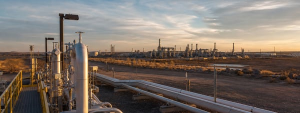 Midstream process piping and a distant oil and gas facility at sunset, used as the feature image for why emissions platforms are replacing disconnected software